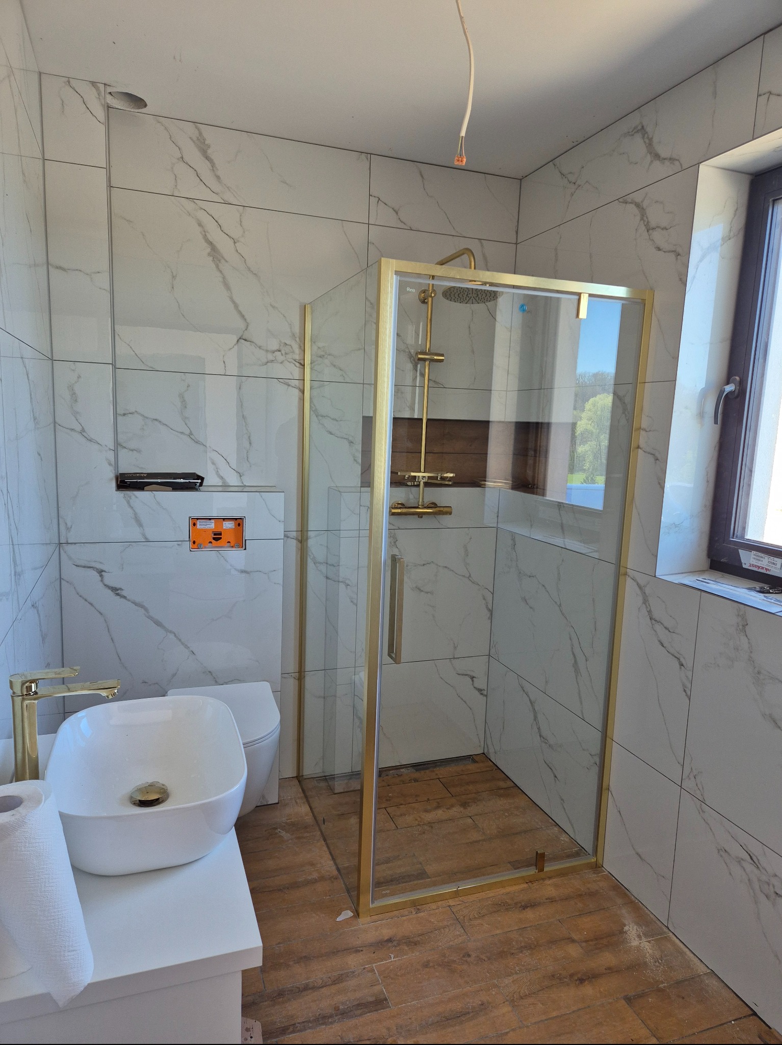 Wykończona łazienka z kabiną prysznicową w złotych ramach, białą umywalką nablatową i toaletą. Ściany wyłożone płytkami imitującymi marmur, drewnopodobna podłoga.