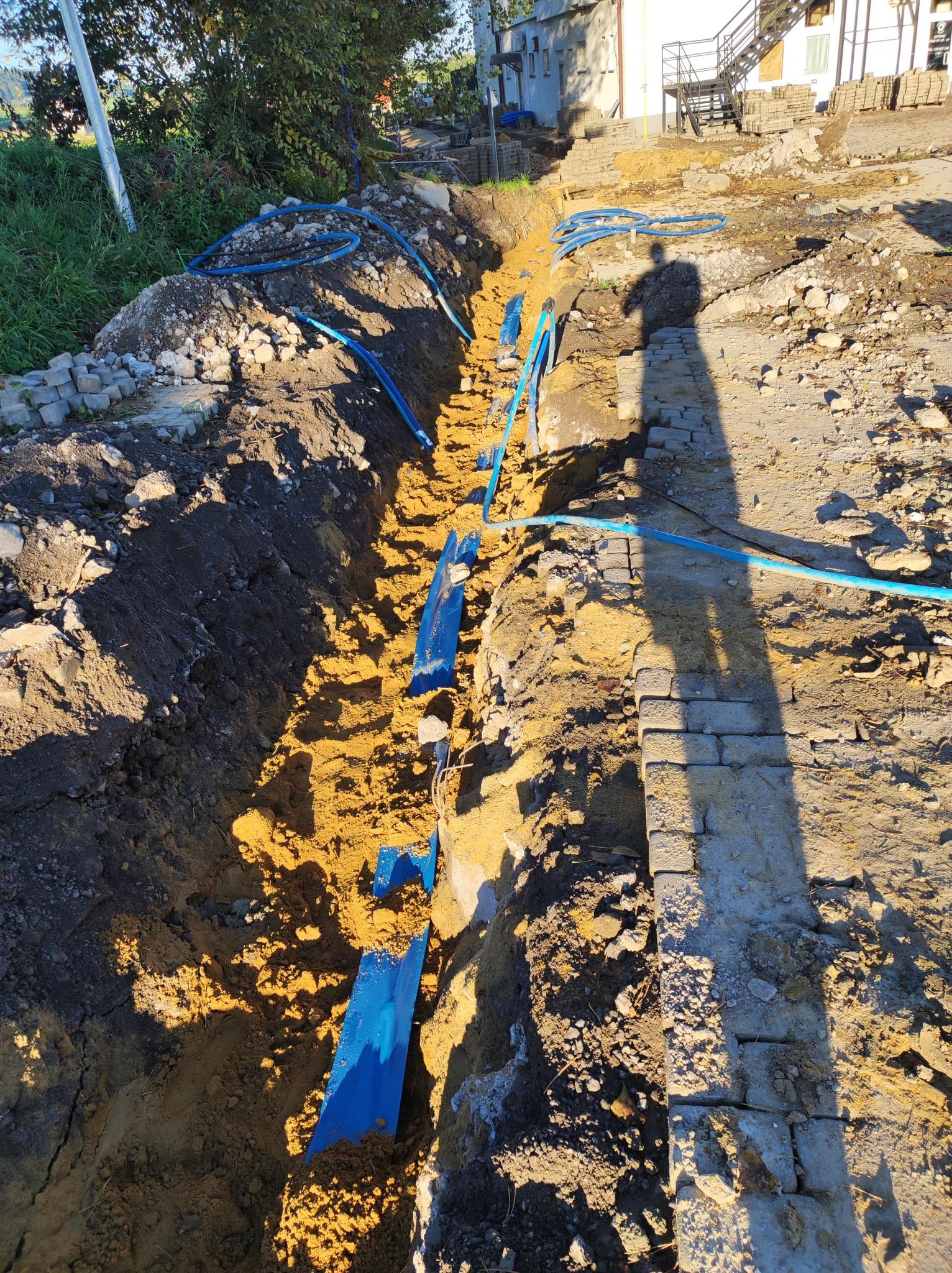 Wykopany rów w ziemi z ułożonymi niebieskimi rurami, w tle widoczny cień osoby i budynek.