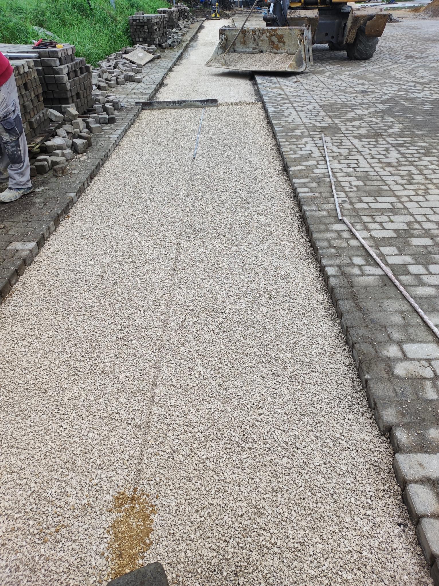 Przygotowanie podłoża z kruszywa pod nawierzchnię z kostki, widoczny równiarka i ułożone obrzeża, w tle stosy kostki brukowej.