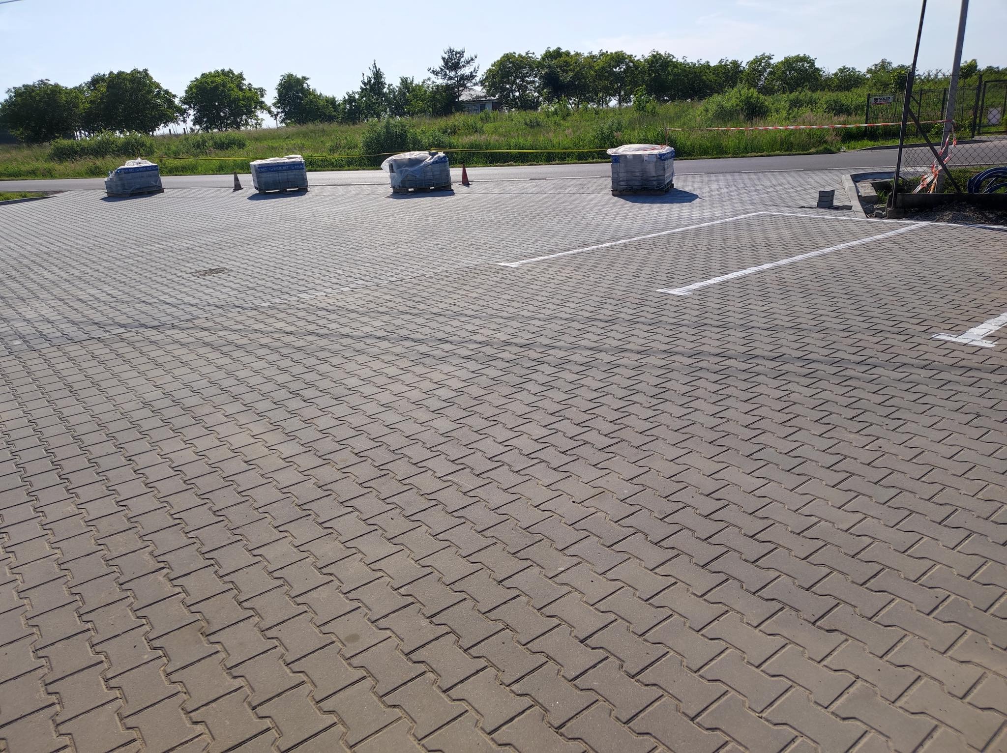 Nowo ułożona, szara kostka brukowa typu 'I', tworząca wzór jodełki na placu parkingowym z wyznaczonymi białymi liniami, palety z kostką w tle, widok z góry pod kątem.