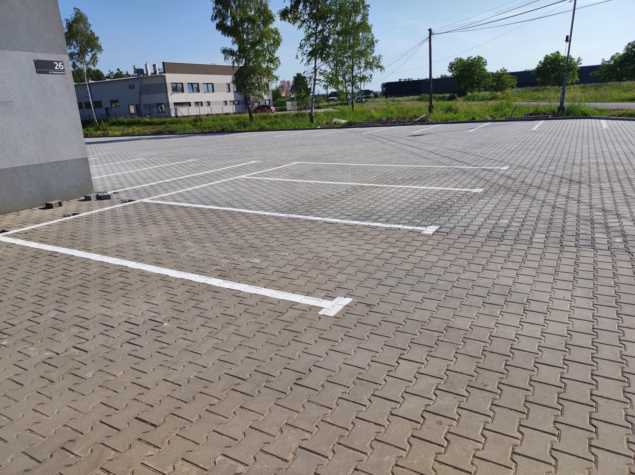 Ułożona kostka brukowa na parkingu z wyznaczonymi białymi liniami miejsc parkingowych, budynek w tle.