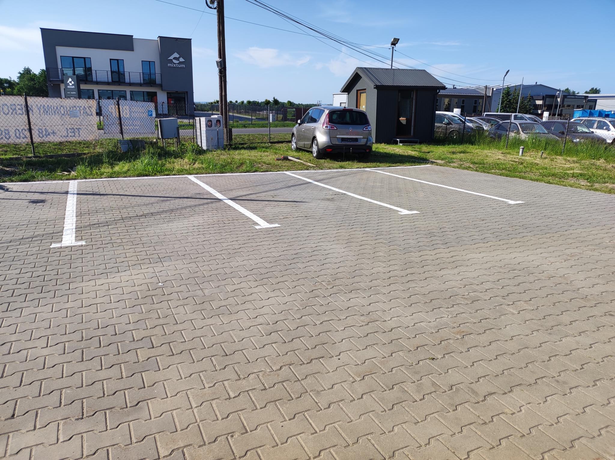 Nowo ułożony parking z szarej kostki brukowej w jodełkę z wyznaczonymi białymi liniami miejsc postojowych, zaparkowany samochód w tle, budynek firmy i inne zaparkowane samochody
