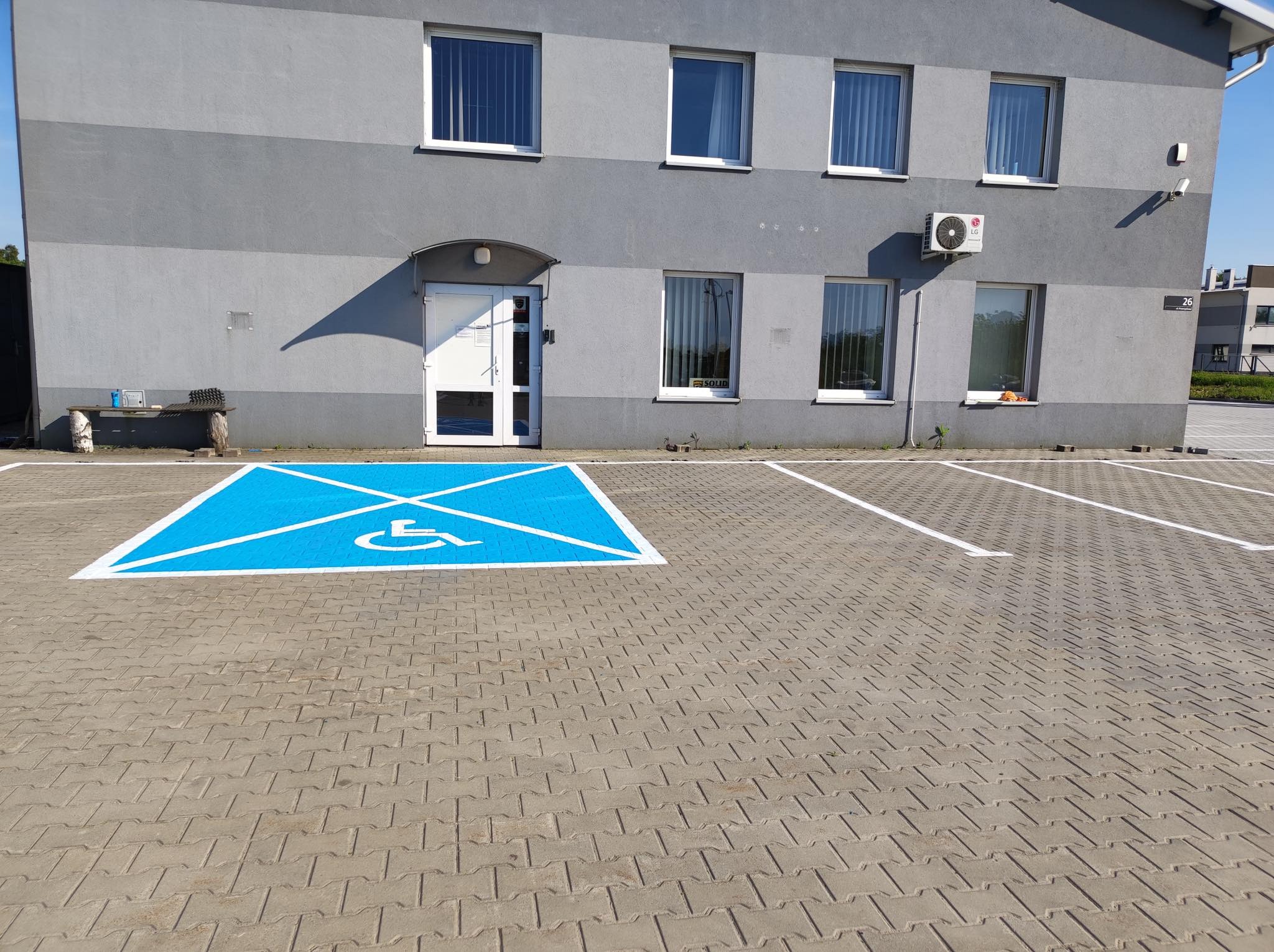 Szary budynek z parkingiem wyłożonym kostką brukową, na którym namalowano niebieskie miejsce dla osoby niepełnosprawnej.