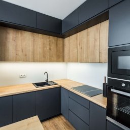 Michał Kopecki - Nowoczesna kuchnia z grafitowymi szafkami dolnymi i drewnopodobnymi szafkami górnymi, czarny zlew i armatura, drewniany blat, sprzęt AGD w zabudowie.