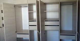 Jasna, drewnopodobna garderoba z otwartymi drzwiami, ukazująca półki, szuflady i drążki na ubrania. Widoczne delikatne, poziome usłojenie na frontach. Minimalistyczny design.