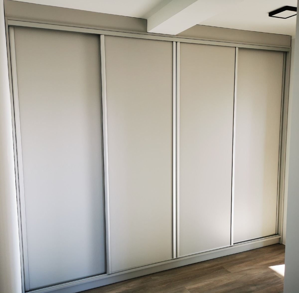 Czterodrzwiowa szafa przesuwna z matowymi, beżowymi frontami i srebrnymi obramowaniami, dopasowana do wnętrza z drewnopodobną podłogą.