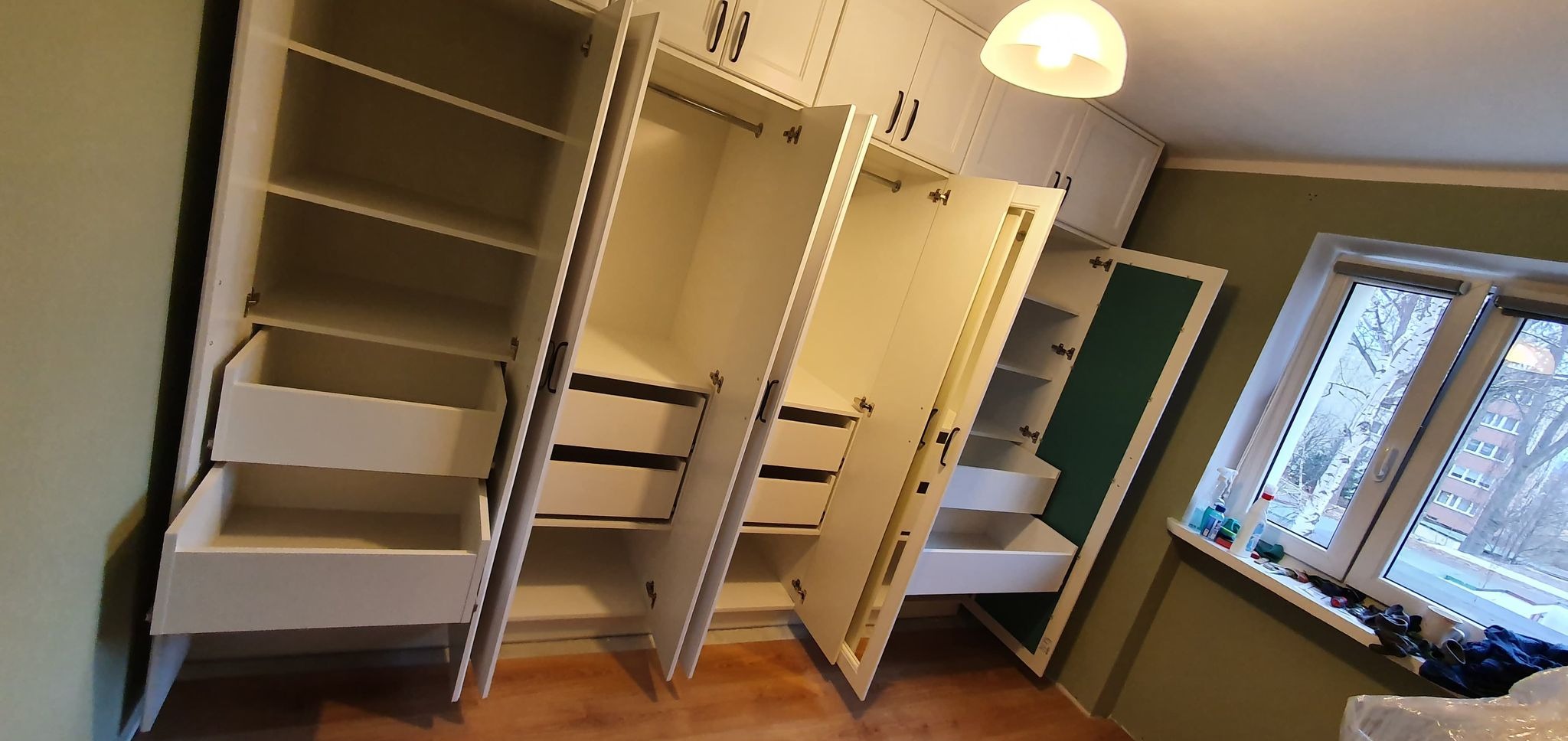 Biała szafa wnękowa z otwartymi drzwiami i szufladami, widok od dołu, w tle okno z widokiem na drzewa.