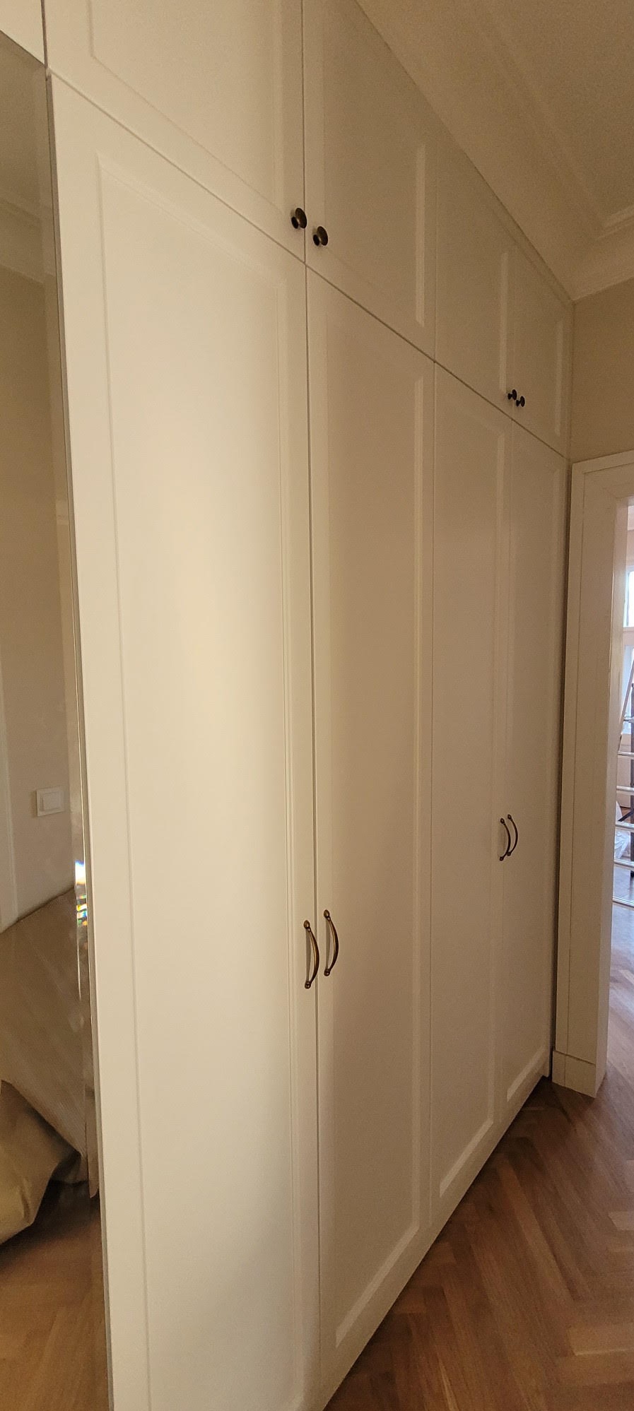 Biała szafa wnękowa z ciemnymi uchwytami, widoczna z boku, z lustrzanymi drzwiami po lewej stronie i parkietem na podłodze.