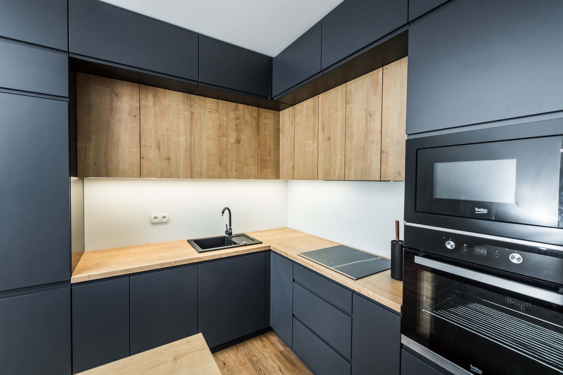 Nowoczesna kuchnia z grafitowymi szafkami dolnymi i drewnopodobnymi szafkami górnymi, czarny zlew i armatura, drewniany blat, sprzęt AGD w zabudowie.