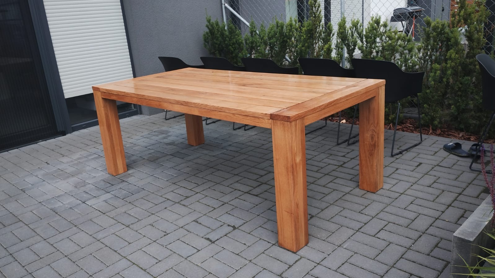 Stół ogrodowy z jasnego drewna o prostokątnym blacie i grubych nogach, ustawiony na szarej kostce brukowej, z czarnymi krzesłami w tle i zielonymi krzewami za nimi.