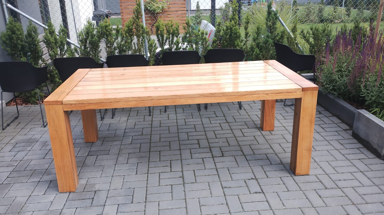 Stół ogrodowy z jasnego drewna o prostej konstrukcji, ustawiony na brukowanej powierzchni, otoczony zielenią i czarnymi krzesłami.