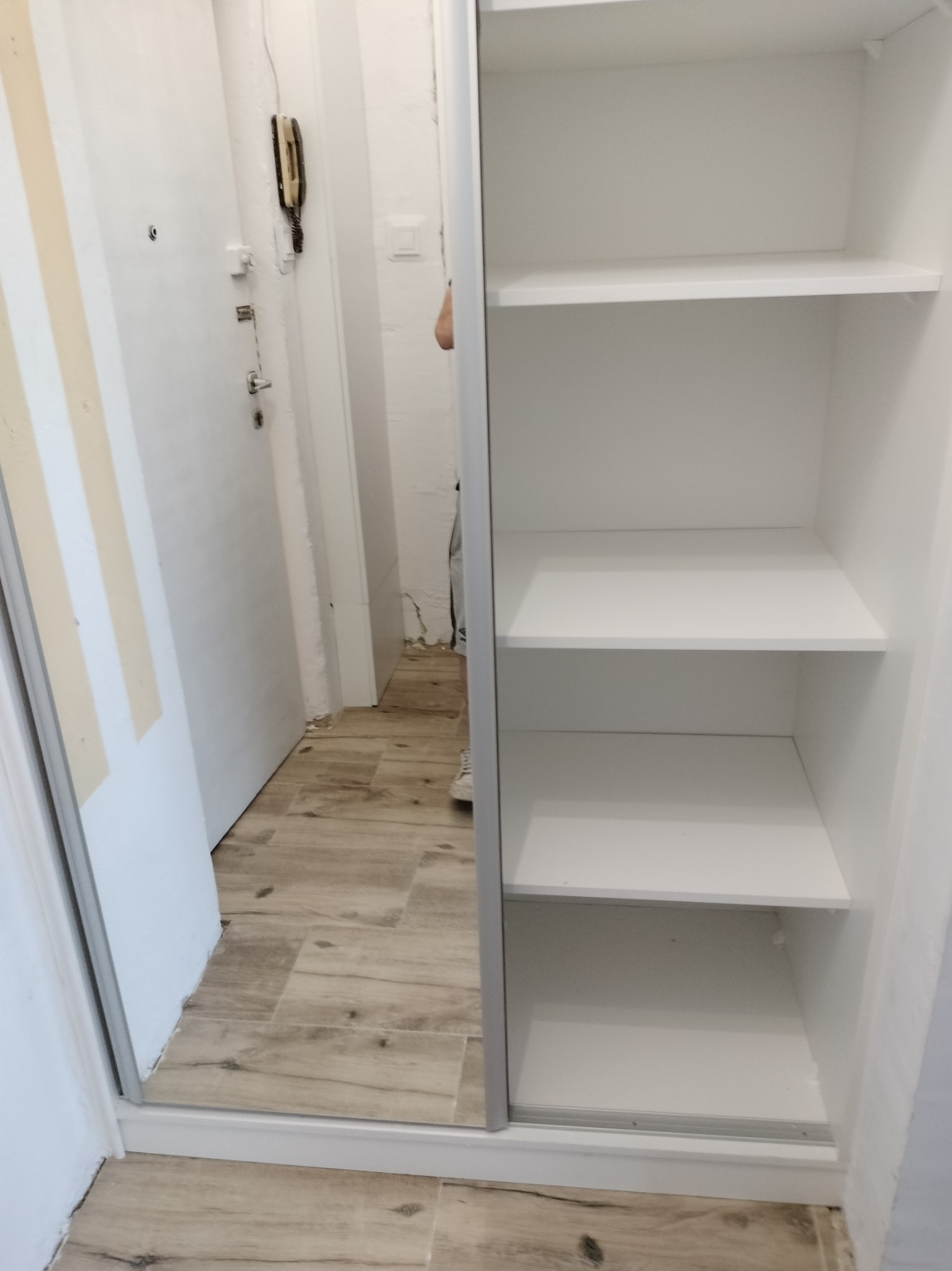 Biała szafa wnękowa z przesuwnymi drzwiami lustrzanymi, widok na wnętrze z półkami i odbicie przedpokoju z drzwiami wejściowymi.