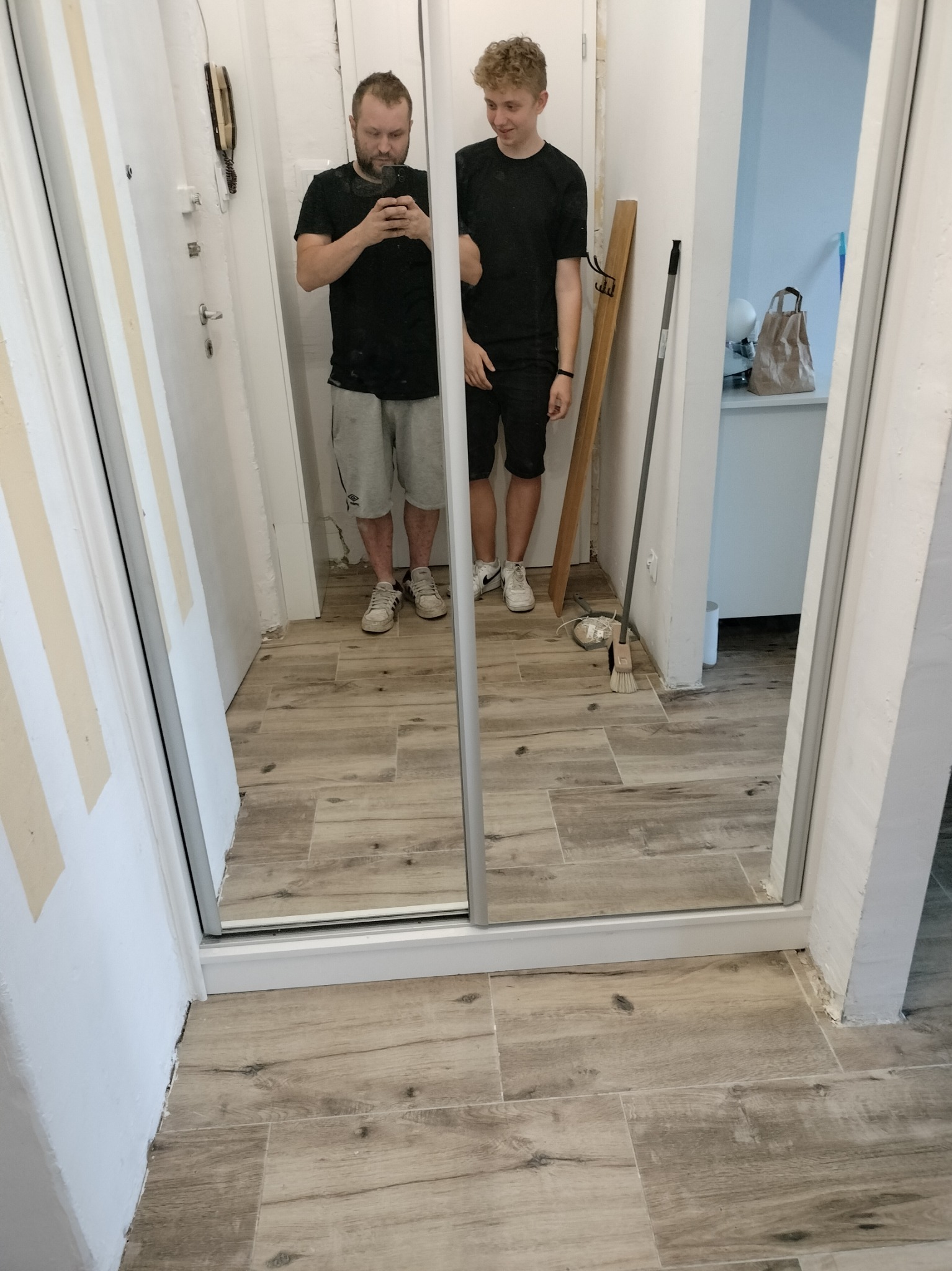 Dwaj mężczyźni widoczni w lustrzanych drzwiach szafy wnękowej, jeden robi zdjęcie telefonem, widoczne niedokończone wykończenie podłogi z paneli imitujących drewno oraz ściany do malowania.