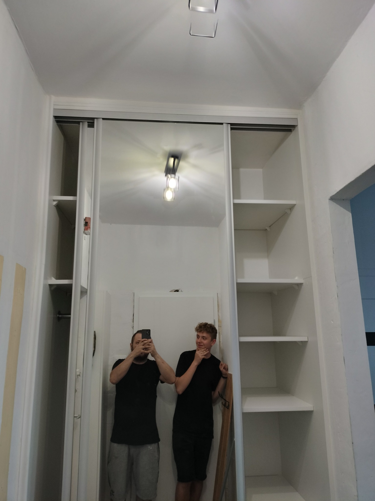 Wnętrze garderoby z przesuwnymi drzwiami, dwie osoby w czarnych koszulkach, jedna robi zdjęcie telefonem, druga zamyślona, białe ściany i półki, minimalistyczne oświetlenie sufitowe.