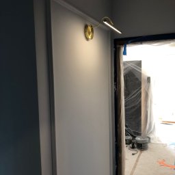 PO MAX - Świeżo pomalowana ściana w ramie z zamontowaną złotą lampką ścienną, widoczne zabezpieczenia folią malarską i pojemniki z farbą w przejściu do kolejnego pomieszczenia.