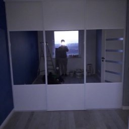 PO MAX - Biała szafa przesuwna z lustrzanymi drzwiami w pokoju z drewnianą podłogą; odbicie osoby robiącej zdjęcie widoczne w lustrze.