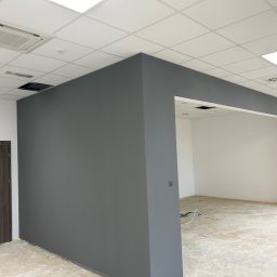 PO MAX - Świeżo pomalowane na szaro ściany biura z widocznym fragmentem sufitu kasetonowego i drzwiami ewakuacyjnymi, na podłodze widoczne ślady po remoncie, w tle mężczyzna w czarnej koszulce.