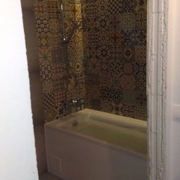 PO MAX - Nowoczesna łazienka z wanną wypełnioną wodą, prysznicem i ścianą wyłożoną wzorzystymi, patchworkowymi płytkami w odcieniach beżu, brązu i niebieskiego, oświetlona listwą LED.