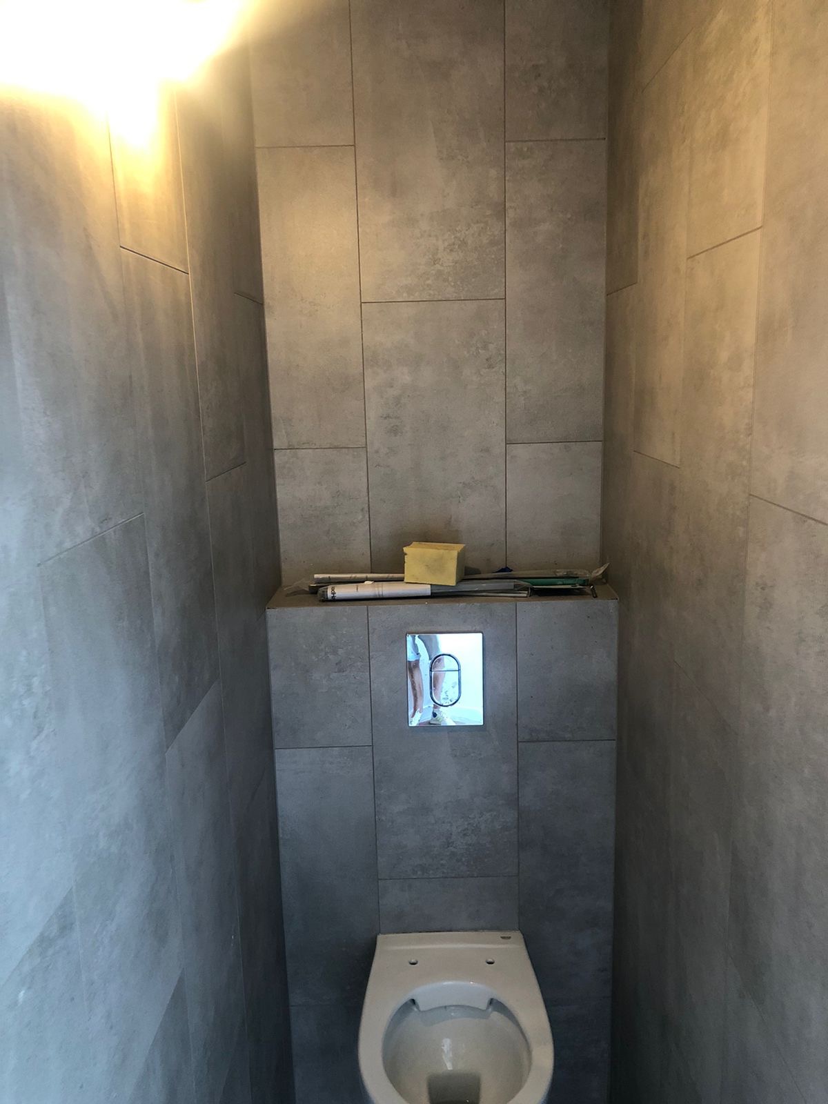 Nowoczesna toaleta w szarej łazience z dużymi płytkami, widoczny przycisk spłuczki i narzędzia na obudowie stelaża podtynkowego.