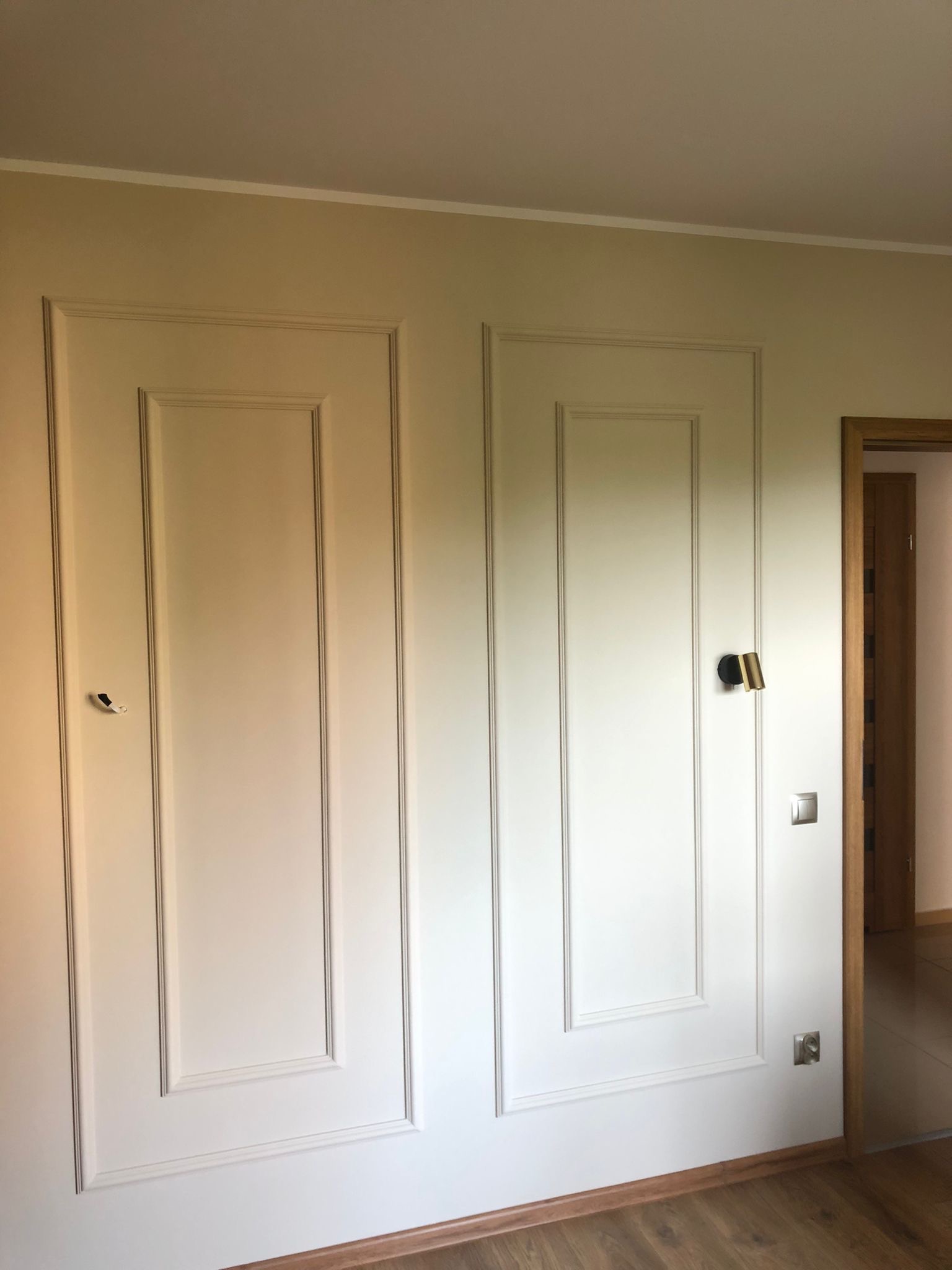 Ściana z dekoracyjnymi panelami w kolorze kremowym, oświetlenie punktowe i widoczny fragment drewnianej podłogi oraz drzwi w tle.