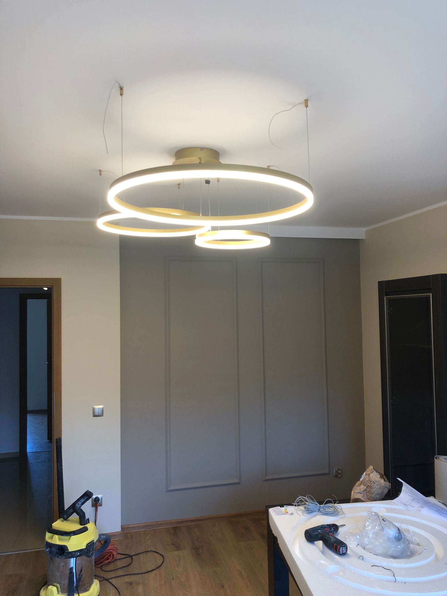 Wnętrze mieszkania w trakcie montażu nowoczesnej, okrągłej lampy LED, widoczny odkurzacz przemysłowy i narzędzia na stole, ściany w kolorze beżowym i szarym z dekoracyjnymi panelami.