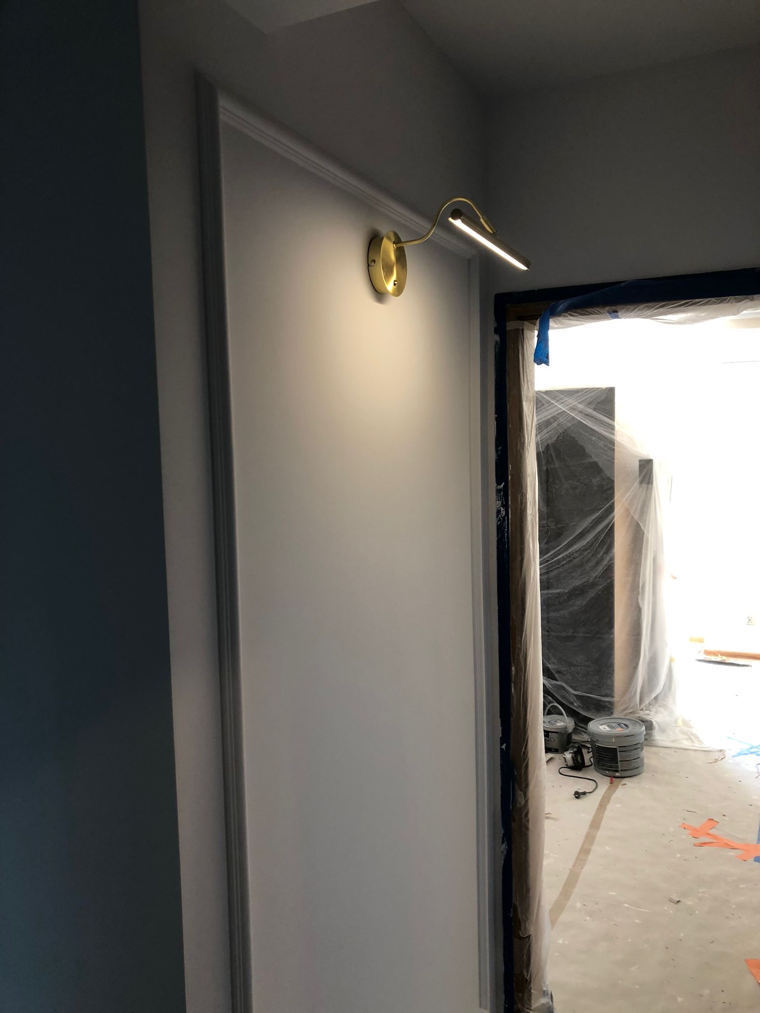 Świeżo pomalowana ściana w ramie z zamontowaną złotą lampką ścienną, widoczne zabezpieczenia folią malarską i pojemniki z farbą w przejściu do kolejnego pomieszczenia.
