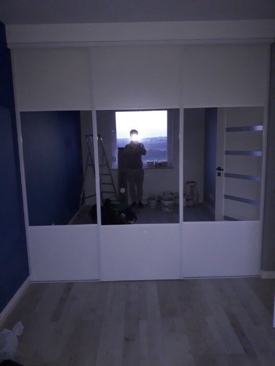 Biała szafa przesuwna z lustrzanymi drzwiami w pokoju z drewnianą podłogą; odbicie osoby robiącej zdjęcie widoczne w lustrze.