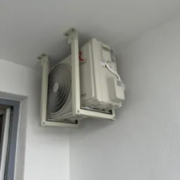 Biały klimatyzator marki Tenso zamontowany na zewnątrz budynku, zawieszony na metalowym stelażu pod sufitem, widok z dołu.