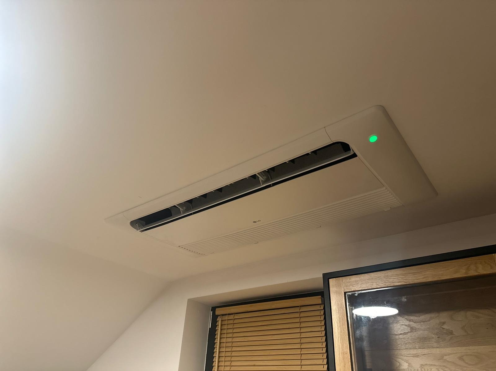 Kasetonowa klimatyzacja LG zamontowana w suficie, widoczna zielona dioda sygnalizacyjna, w tle okno z żaluzjami i fragment drewnianej ramy lustra.