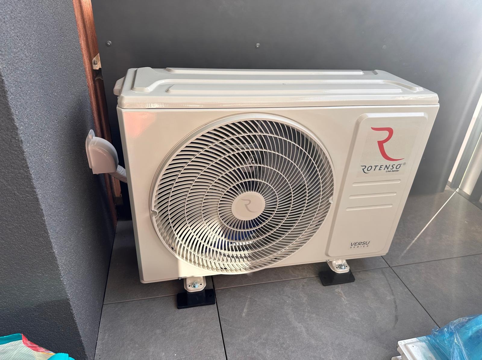 Zewnętrzna jednostka klimatyzacji Rotenso Versu Series, zamontowana na czarnych podkładkach, widoczna przy szarej ścianie, z widocznym przewodem w osłonie.