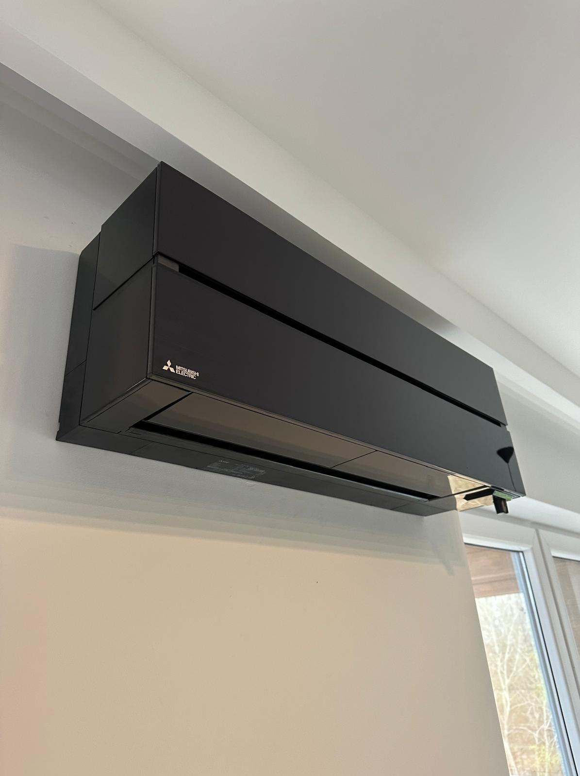 Czarna jednostka klimatyzacji Mitsubishi Electric zamontowana na ścianie w jasnym pomieszczeniu, widok z dołu, okno w tle.
