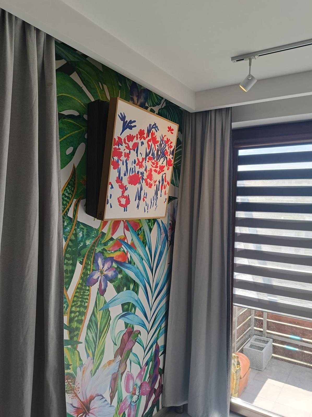 Klimatyzator ścienny w obudowie z drewnianą ramą, wkomponowany w tropikalną tapetę, obok szarych zasłon i okna z żaluzjami.