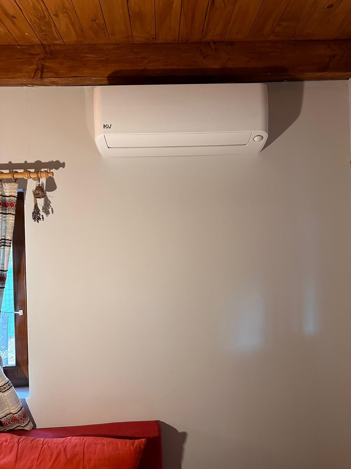 Biały klimatyzator na ścianie w pokoju z drewnianym sufitem i oknem z ozdobnymi zasłonami.