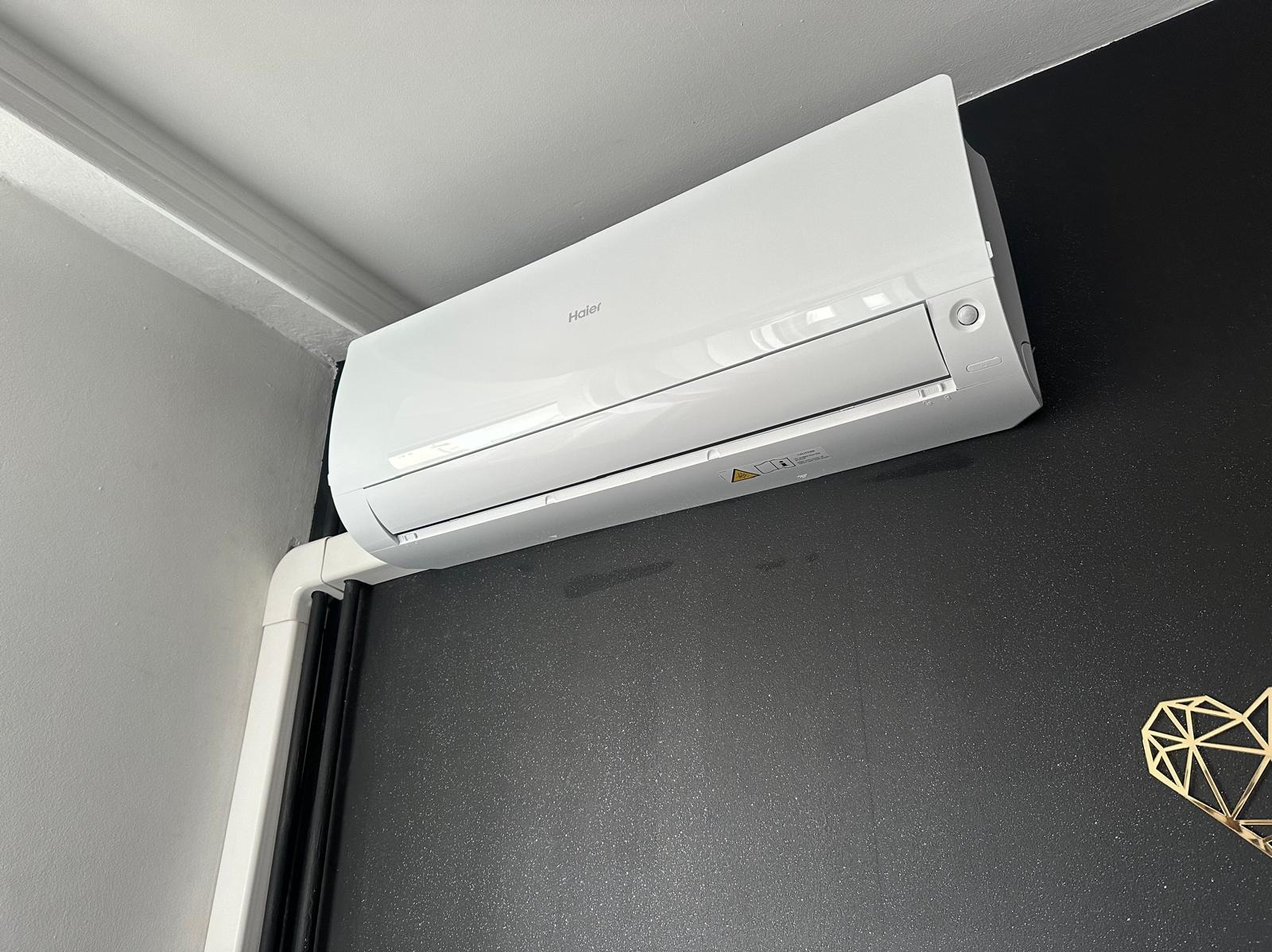 Biała klimatyzacja Haier zamontowana na ciemnoszarej ścianie z dekoracją w kształcie serca, widoczne rury instalacyjne