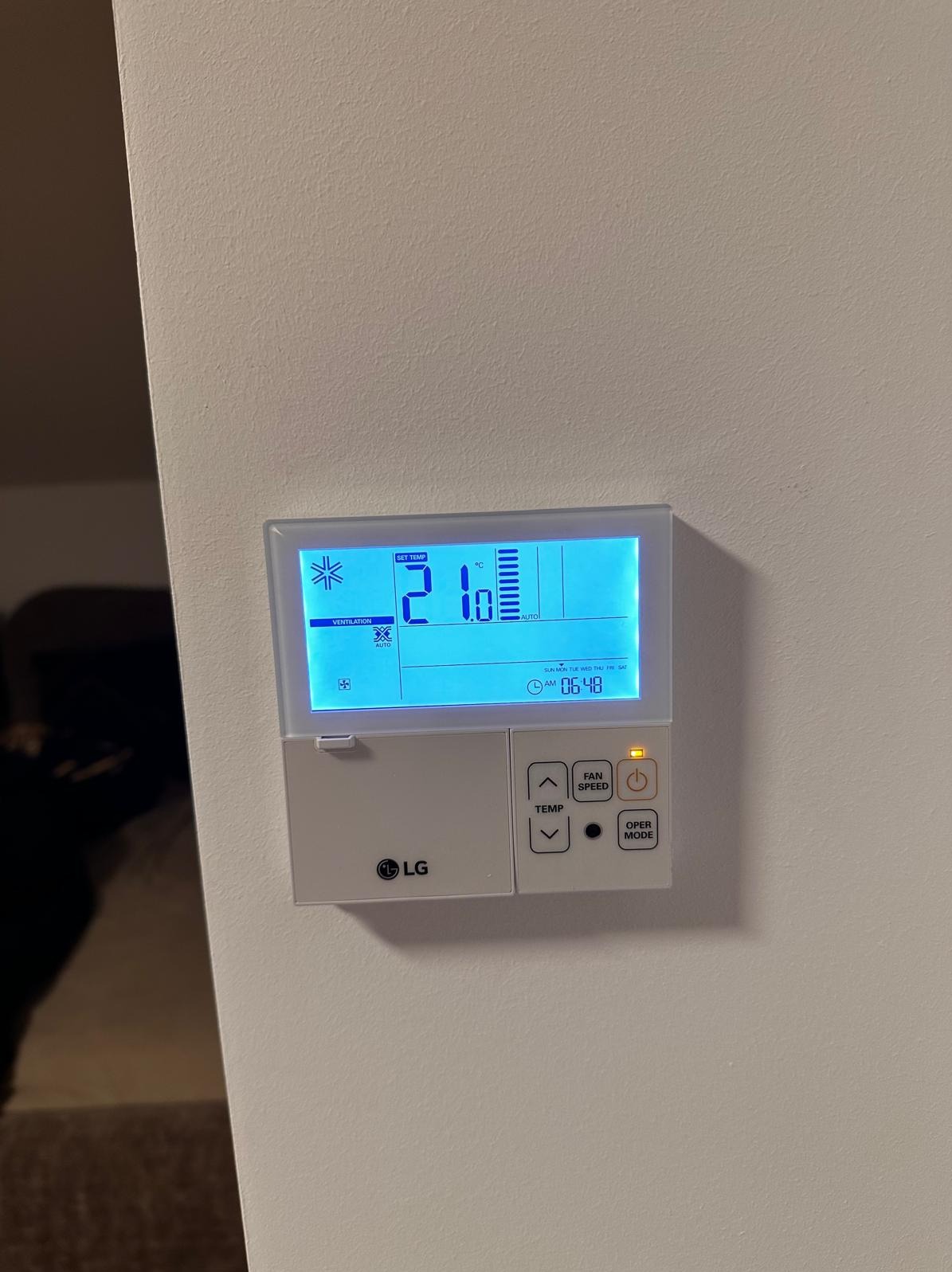 Biały panel sterowania LG klimatyzacji z wyświetlaczem LCD wskazującym temperaturę 21 stopni Celsjusza, ustawienia wentylacji i tryb automatyczny, zamontowany na jasnej ścianie.