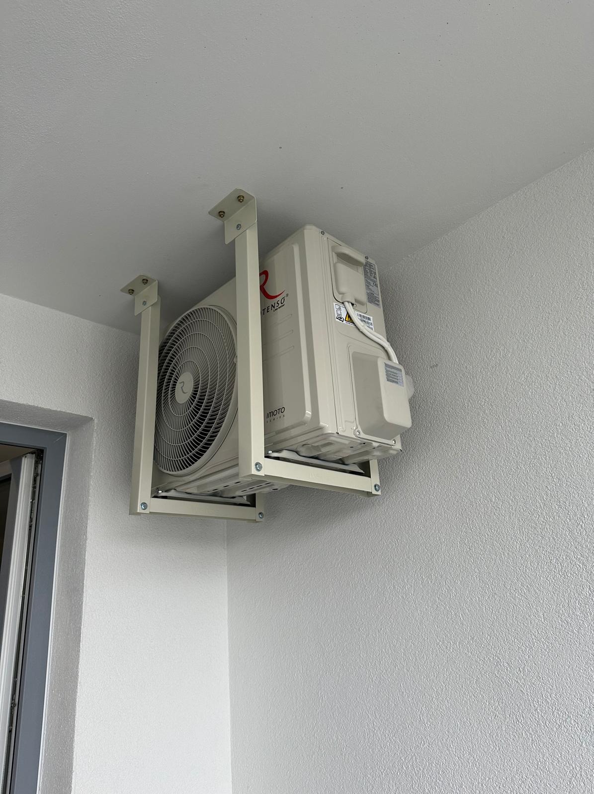 Biały klimatyzator marki Tenso zamontowany na zewnątrz budynku, zawieszony na metalowym stelażu pod sufitem, widok z dołu.