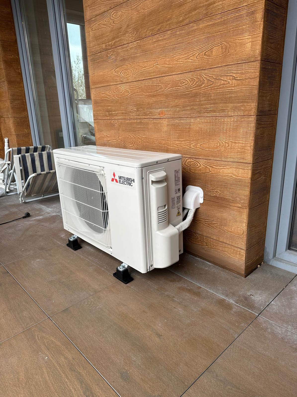 Biała, zewnętrzna jednostka klimatyzacji Mitsubishi Electric na czarnych podstawkach, zamontowana na brązowej, drewnopodobnej elewacji balkonu.