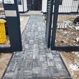 Spaw-Bud Klaudia Bartoszewska - Nowo ułożona, szara kostka brukowa prowadząca od furtki z metalowymi słupkami i numerem domu do drzwi wejściowych, widoczna w zimowej scenerii z resztkami śniegu.