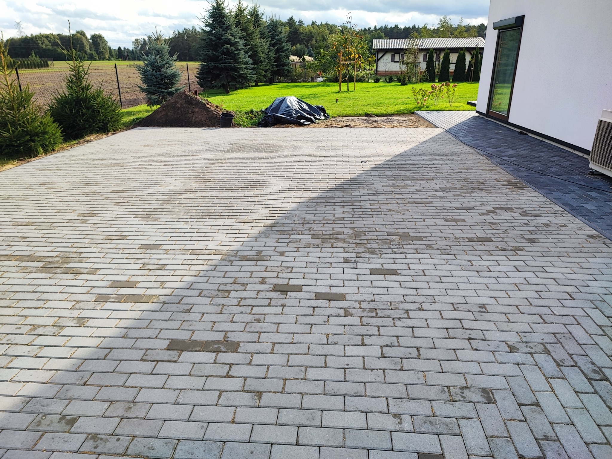 Nowo ułożona, szara kostka brukowa w jodełkę na podjeździe przed domem, z widocznym stosem ziemi i czarną plandeką w tle, kontrastująca z ciemniejszą kostką przy ścianie budynku.