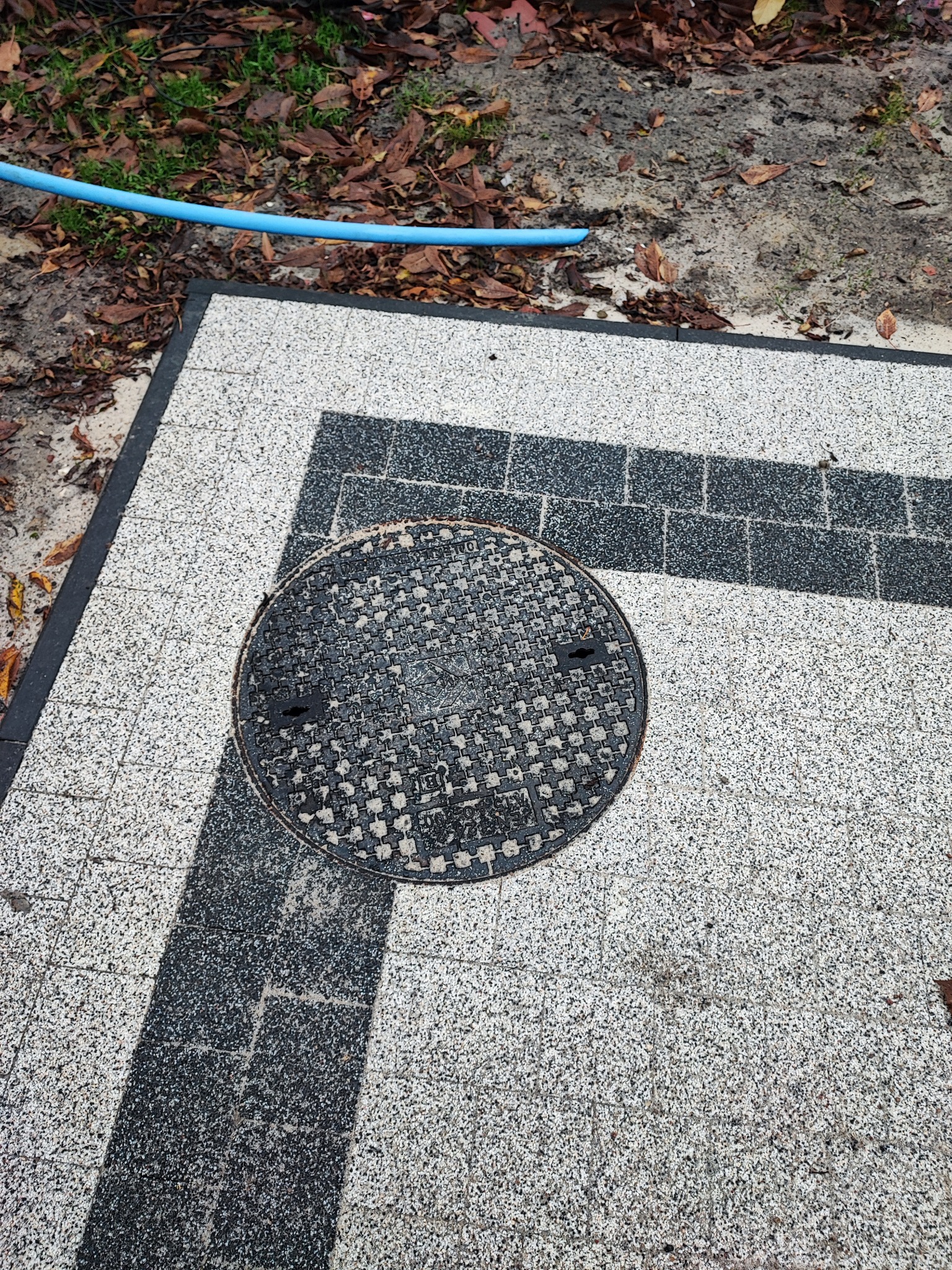 Szara kostka brukowa z ciemnym wzorem i okrągła studzienka kanalizacyjna, obok leży niebieski wąż ogrodowy.