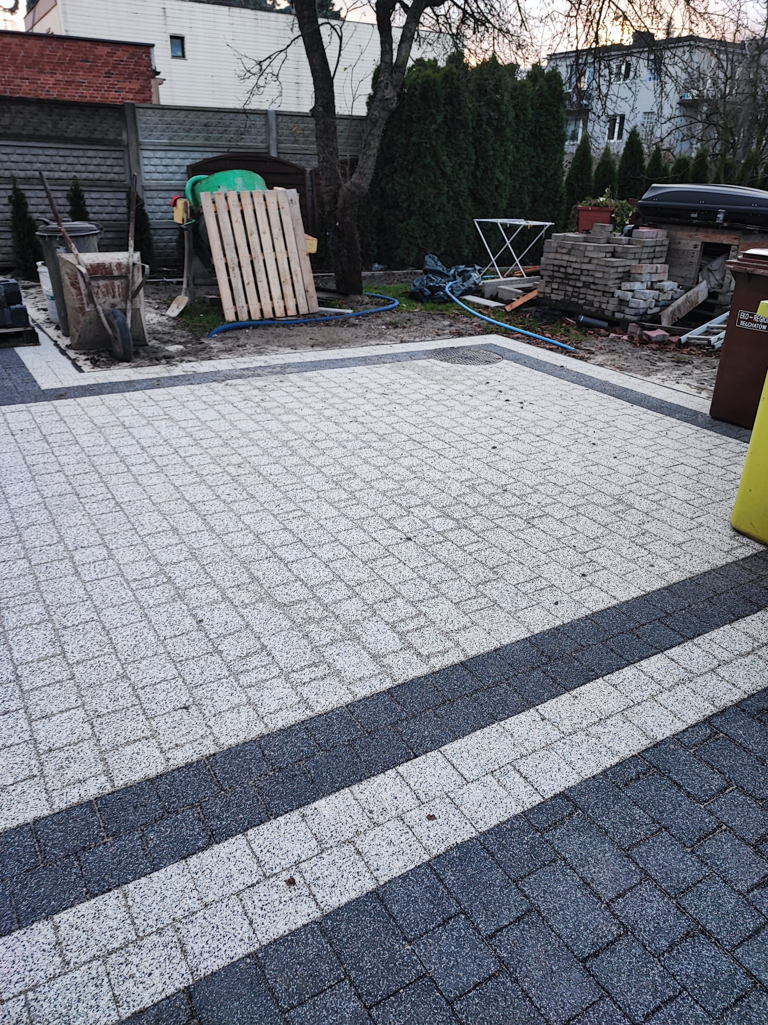 Ułożona nawierzchnia z kostki brukowej w odcieniach szarości i bieli, z ciemniejszymi pasami, widoczne narzędzia i materiały budowlane w tle.