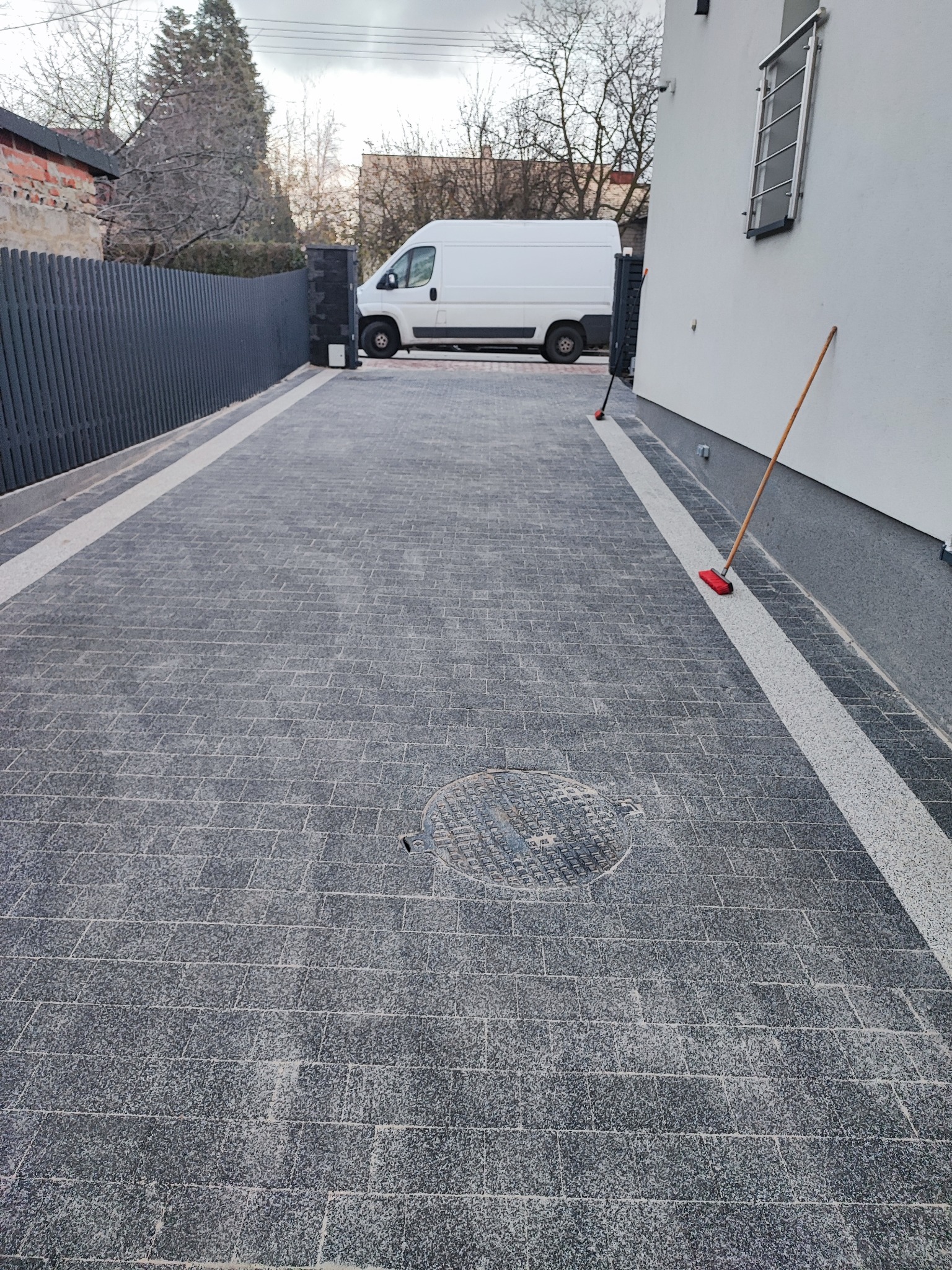 Nowo ułożona, szara kostka granitowa z jasnymi pasami po bokach, studzienka kanalizacyjna na środku, szara brama w tle, biały bus i ściana budynku.