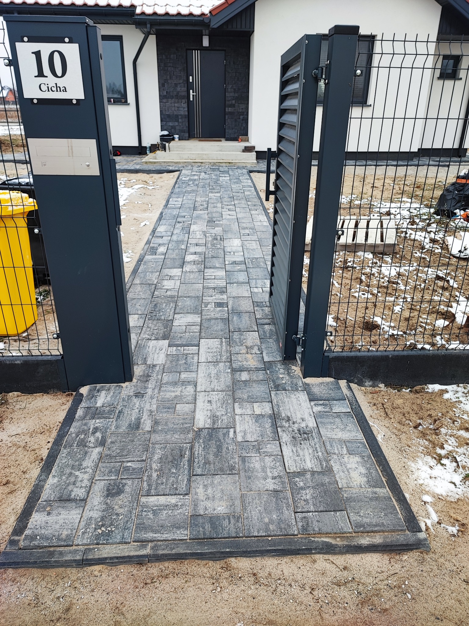 Nowo ułożona, szara kostka brukowa prowadząca od furtki z metalowymi słupkami i numerem domu do drzwi wejściowych, widoczna w zimowej scenerii z resztkami śniegu.