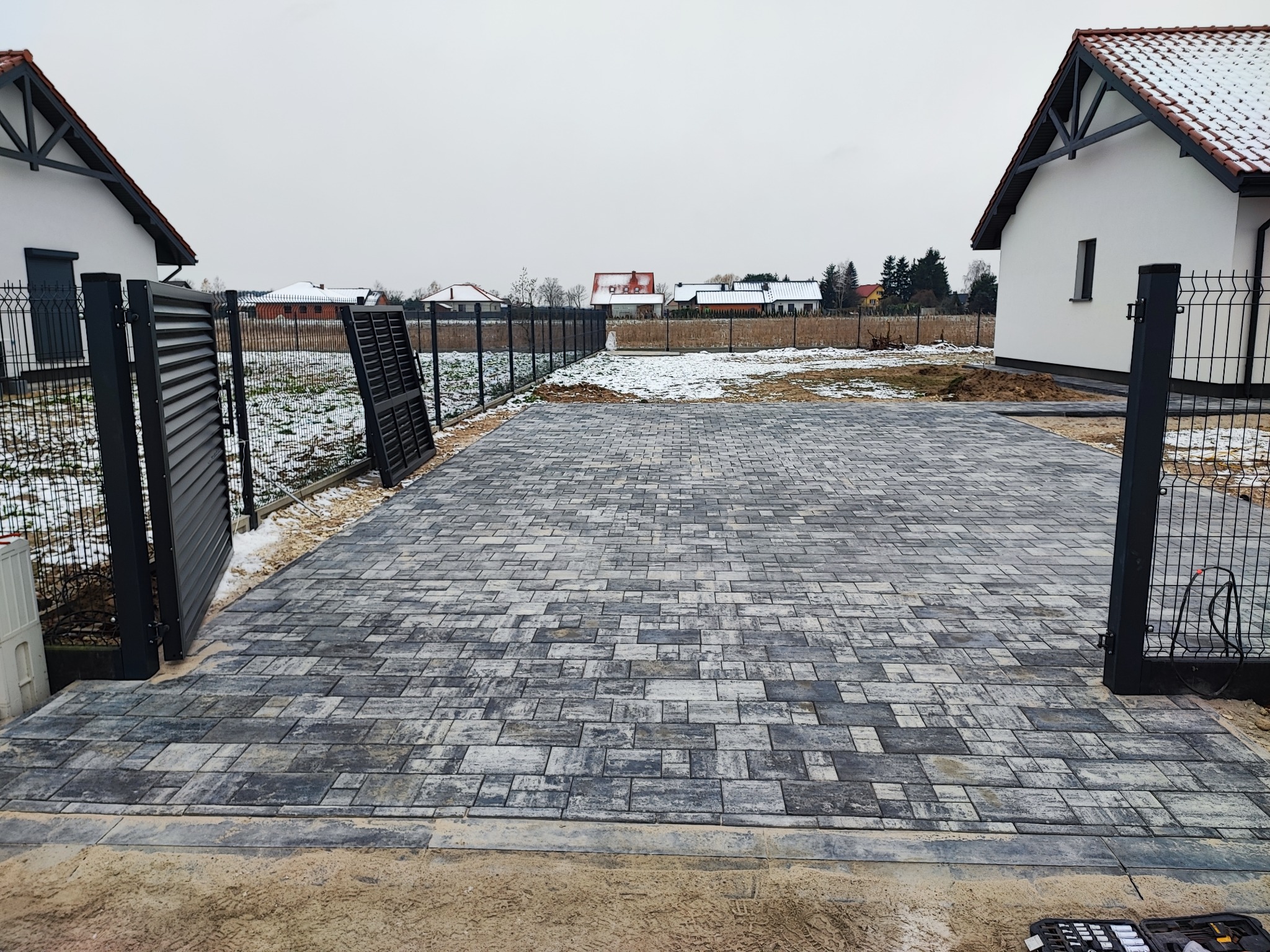 Ułożona szara kostka brukowa na podjeździe między dwoma domami, widoczna brama wjazdowa i narzędzia brukarza w prawym dolnym rogu, w tle ogrodzenie i częściowo ośnieżony teren.