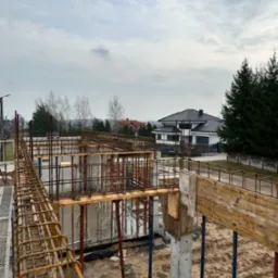 Wiązanie zbrojenia na fundamencie domu jednorodzinnego przed wylaniem betonu, widoczne szalunki i podpory, w tle dom w stanie surowym zamkniętym.