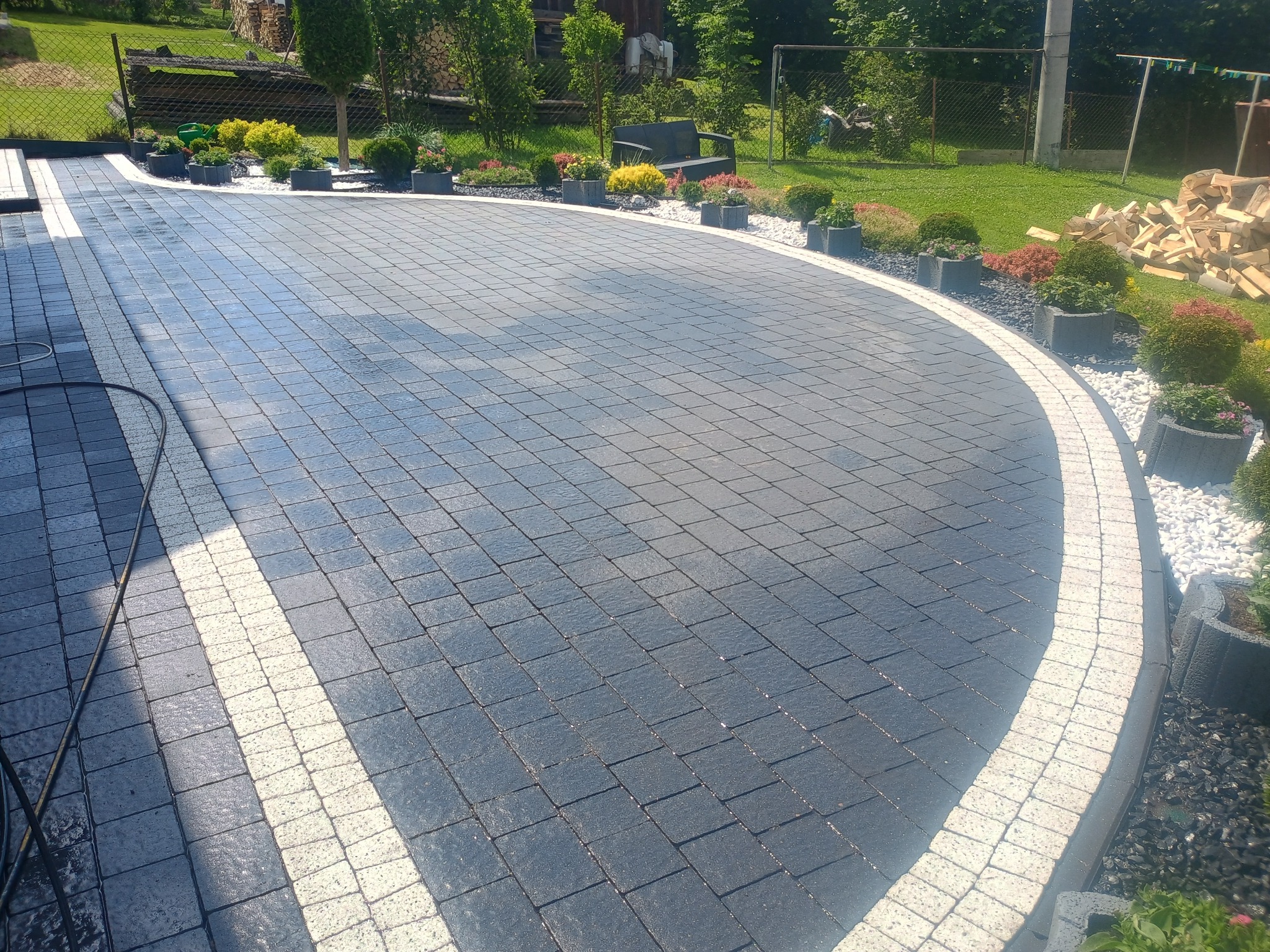 Ułożona z ciemnej kostki brukowej powierzchnia z białymi obrzeżami, otoczona rabatami z kwiatami i ozdobnymi kamieniami, widoczny wąż ogrodowy.