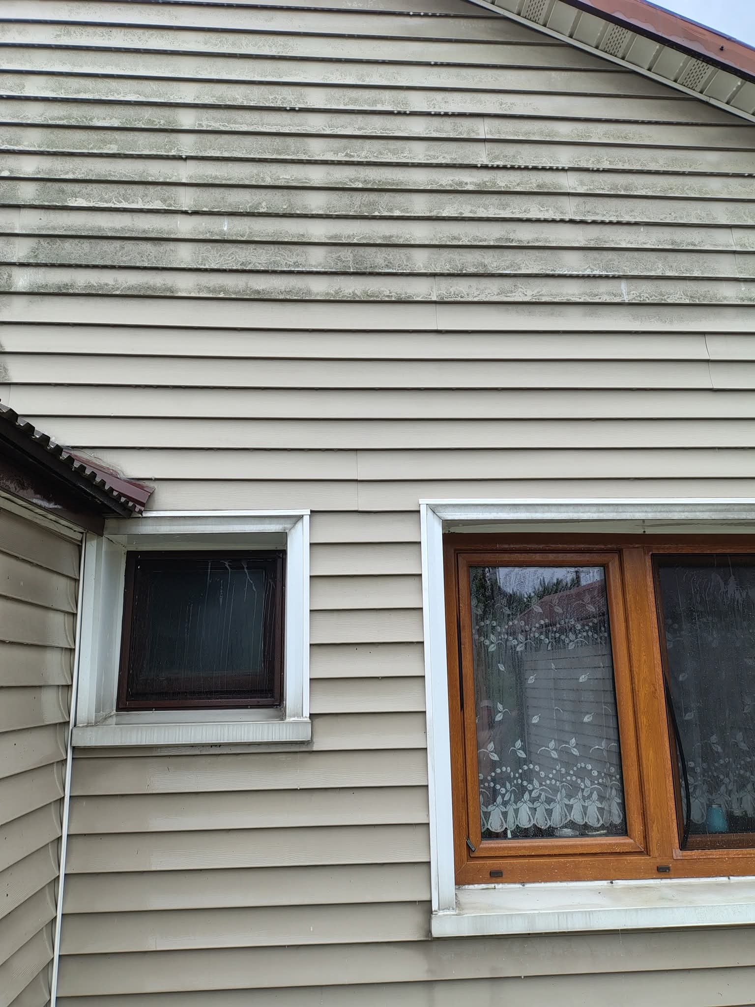 Zabrudzona elewacja domu z sidingu, widoczne okna z drewnianą ramą. Szare zabrudzenia na jasnym tle sidingu. Ujęcie z dołu, fragment dachu.