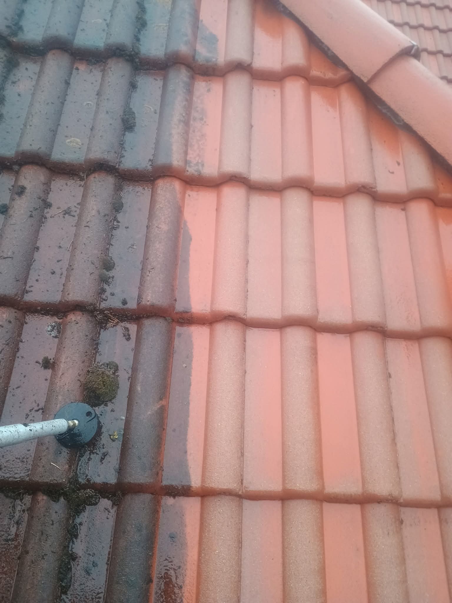 Częściowo umyta dachówka ceramiczna, widoczna różnica między brudną, porośniętą mchem powierzchnią a czystą, czerwoną dachówką po myciu. Dysza myjąca w lewym dolnym rogu.
