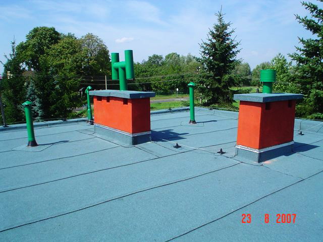 Dwa pomarańczowe kominy z szarymi daszkami i zielonymi rurami wentylacyjnymi na płaskim, bitumicznym dachu w słoneczny dzień.