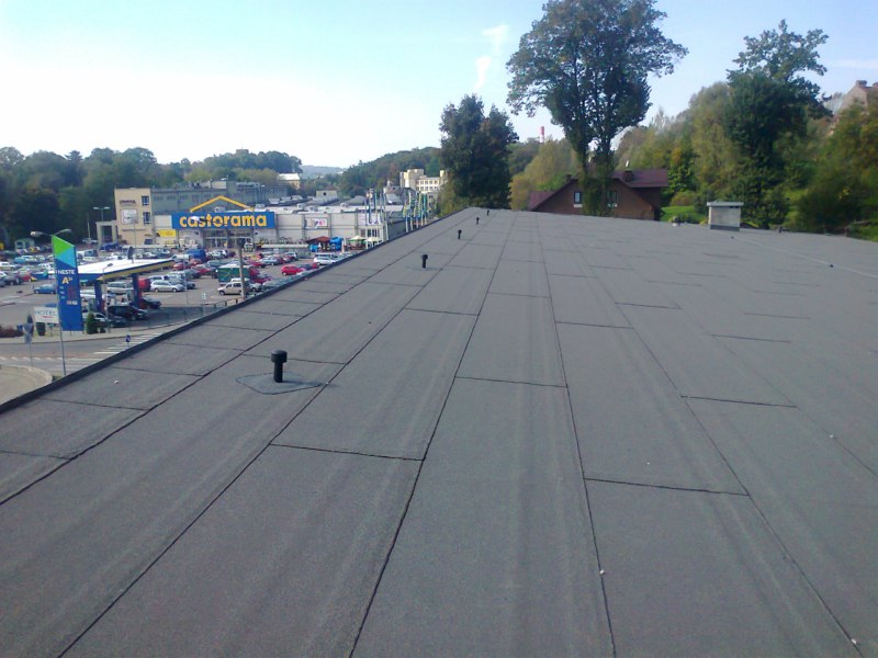 Czarny, płaski dach pokryty papą termozgrzewalną na tle parkingu sklepu Castorama i zieleni drzew.